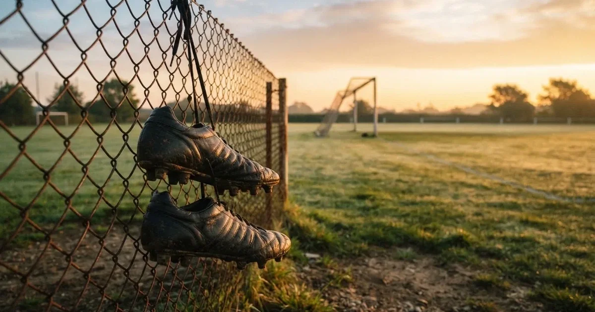 Znoszone buty piłkarskie zawieszone za sznurówki na drucianym płocie na skraju boiska treningowego o wschodzie słońca.