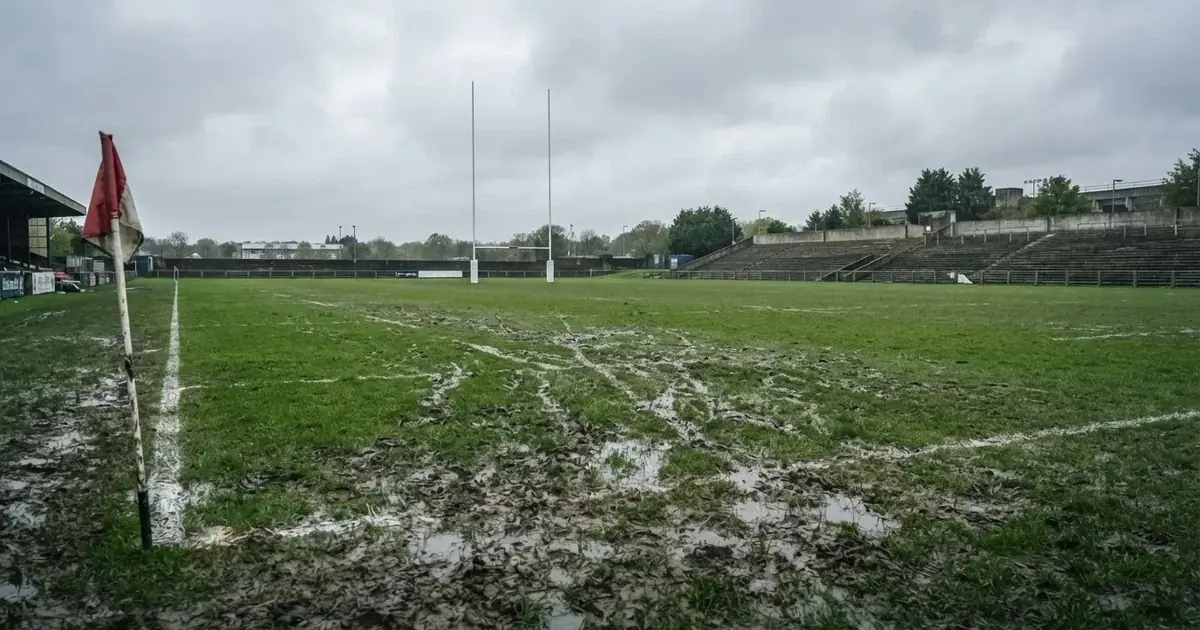 Boisko do rugby ze słupami bramki w oddali pod pochmurnym niebem, mokra trawa i ślady butów.